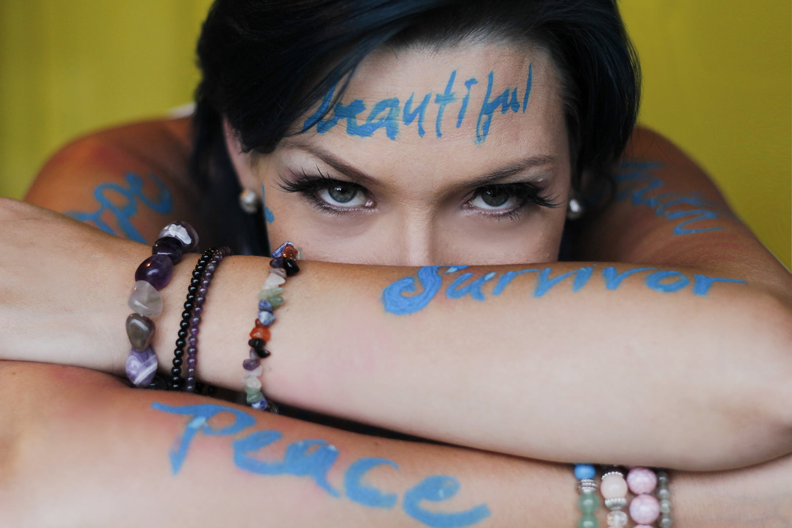 Peaceful Anna looks forward. Blue words 'beautiful,' 'peace,' and 'survivor' are painted on arms and face.