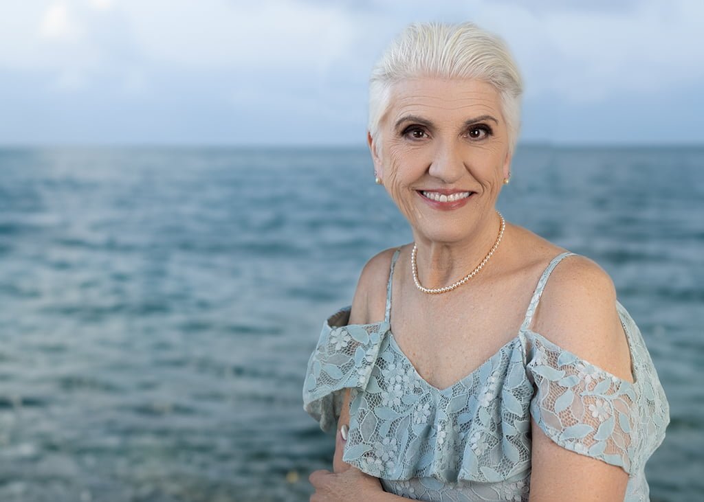 Sharon has lacy blue dress and smiles with pearl earrings and a necklace. She is in front of a blue ocean. Her fingernails are painted platinum.