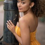 Gallery image of lack Model with curly Hair and a mustard yellow striped dress standing.