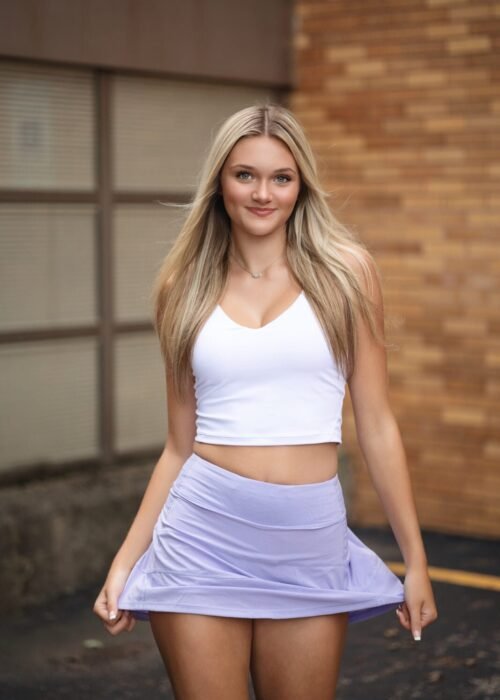 Model with blonde hair walking forward. She is touching her hem of her lavender skirt and has a white top on.