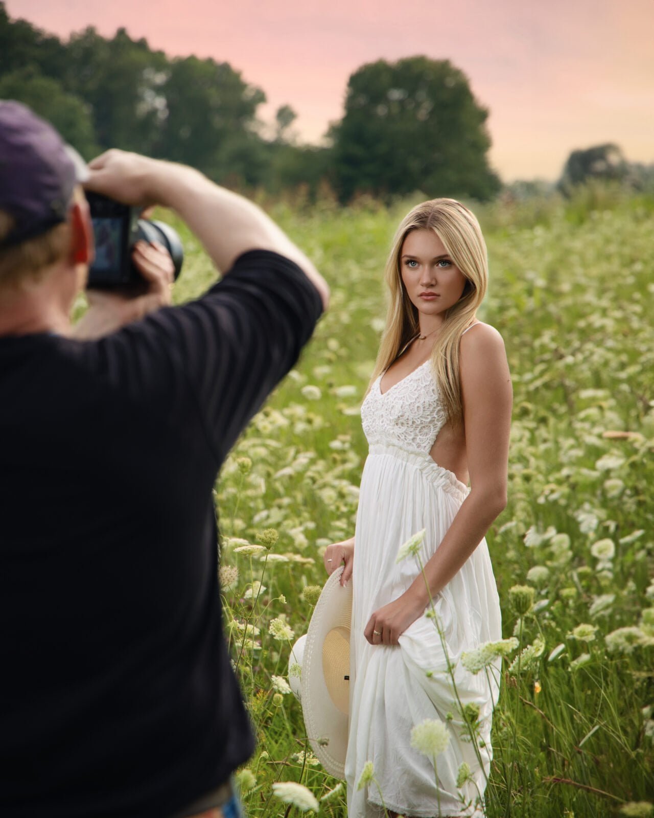 A man in a hat and a black shirt is taking a picture of a girl in a field of wild white flowers in a white dress.