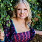 Gallery image of A high school senior from Granville High School gently holds a branch of lush green ivy on the wall by her pool, smiling warmly. This is her outdoor senior photography session. Wearing a vibrant, bohemian-style floral dress in deep purple, pink, and blue, she blends seamlessly with the ivy-covered background. She has layered gold necklaces and hoop earrings. Desired Focus Photography specializes in senior portraits that capture each student's individuality, creating timeless memories of this milestone year in a natural, outdoor setting. Perfect for families looking for professional senior photography that highlights authenticity and beauty.