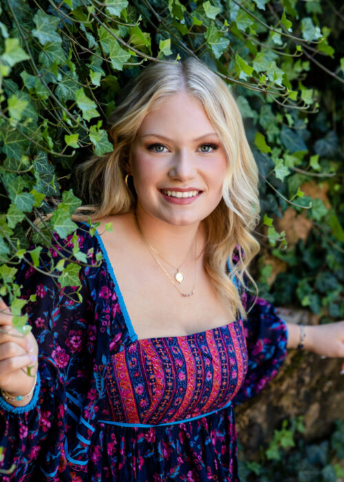 A high school senior from Granville High School gently holds a branch of lush green ivy on the wall by her pool, smiling warmly. This is her outdoor senior photography session. Wearing a vibrant, bohemian-style floral dress in deep purple, pink, and blue, she blends seamlessly with the ivy-covered background. She has layered gold necklaces and hoop earrings. Desired Focus Photography specializes in senior portraits that capture each student's individuality, creating timeless memories of this milestone year in a natural, outdoor setting. Perfect for families looking for professional senior photography that highlights authenticity and beauty.