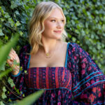 Gallery image of Here's another image we loved. It would be perfect for a cover of a magazine. A high school senior from Granville High School stands gracefully in front of a wall of lush green ivy, with her hand gently resting on the leaves. She gazes off to the side with a soft, confident smile, capturing a moment of natural beauty. Her blonde hair frames her face, and she wears a vibrant, bohemian-style floral dress in shades of deep purple, pink, and blue, complemented by delicate gold necklaces. This image, taken by Desired Focus Photography.