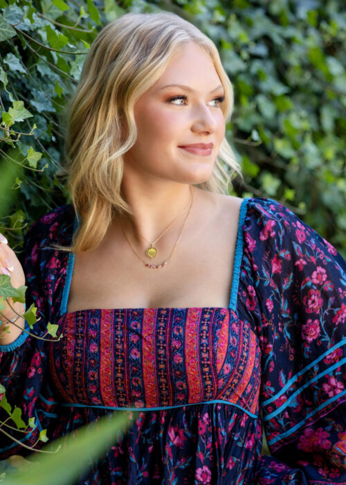 Here's another image we loved. It would be perfect for a cover of a magazine. A high school senior from Granville High School stands gracefully in front of a wall of lush green ivy, with her hand gently resting on the leaves. She gazes off to the side with a soft, confident smile, capturing a moment of natural beauty. Her blonde hair frames her face, and she wears a vibrant, bohemian-style floral dress in shades of deep purple, pink, and blue, complemented by delicate gold necklaces. This image, taken by Desired Focus Photography.